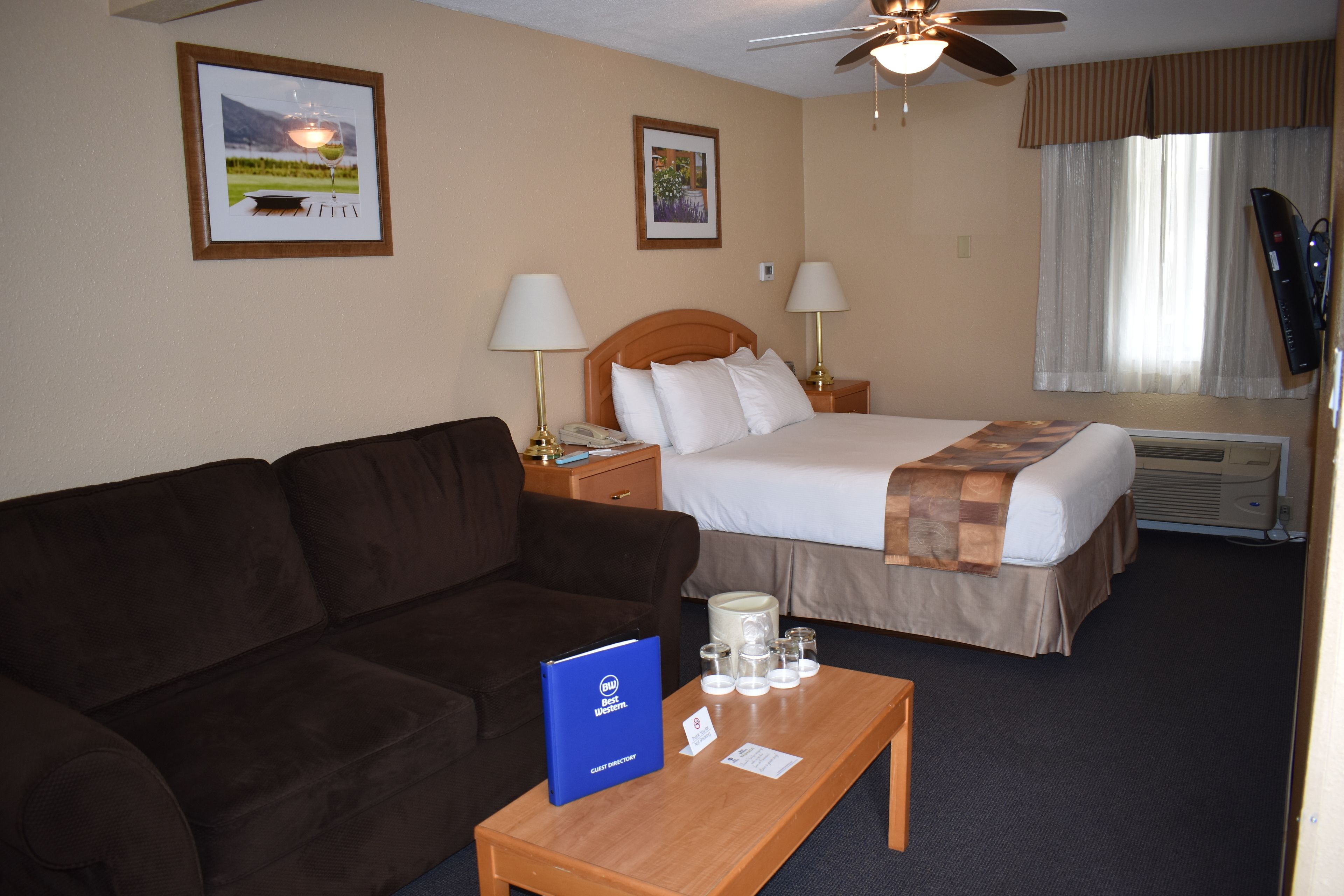 Habitación de hotel con cama, sofá, mesita de noche con lámpara, ventilador de techo y cuadros en la pared.