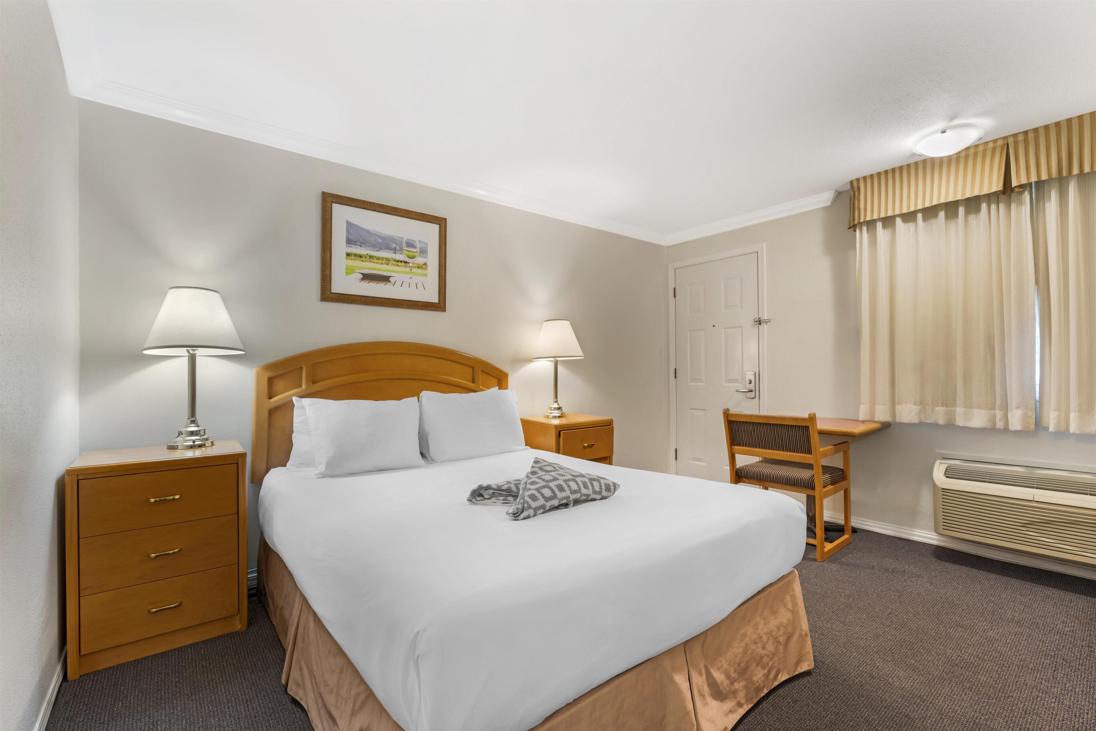 Habitación de hotel con cama, dos mesitas de noche, una silla, un escritorio, un cuadro en la pared y una ventana con cortinas.