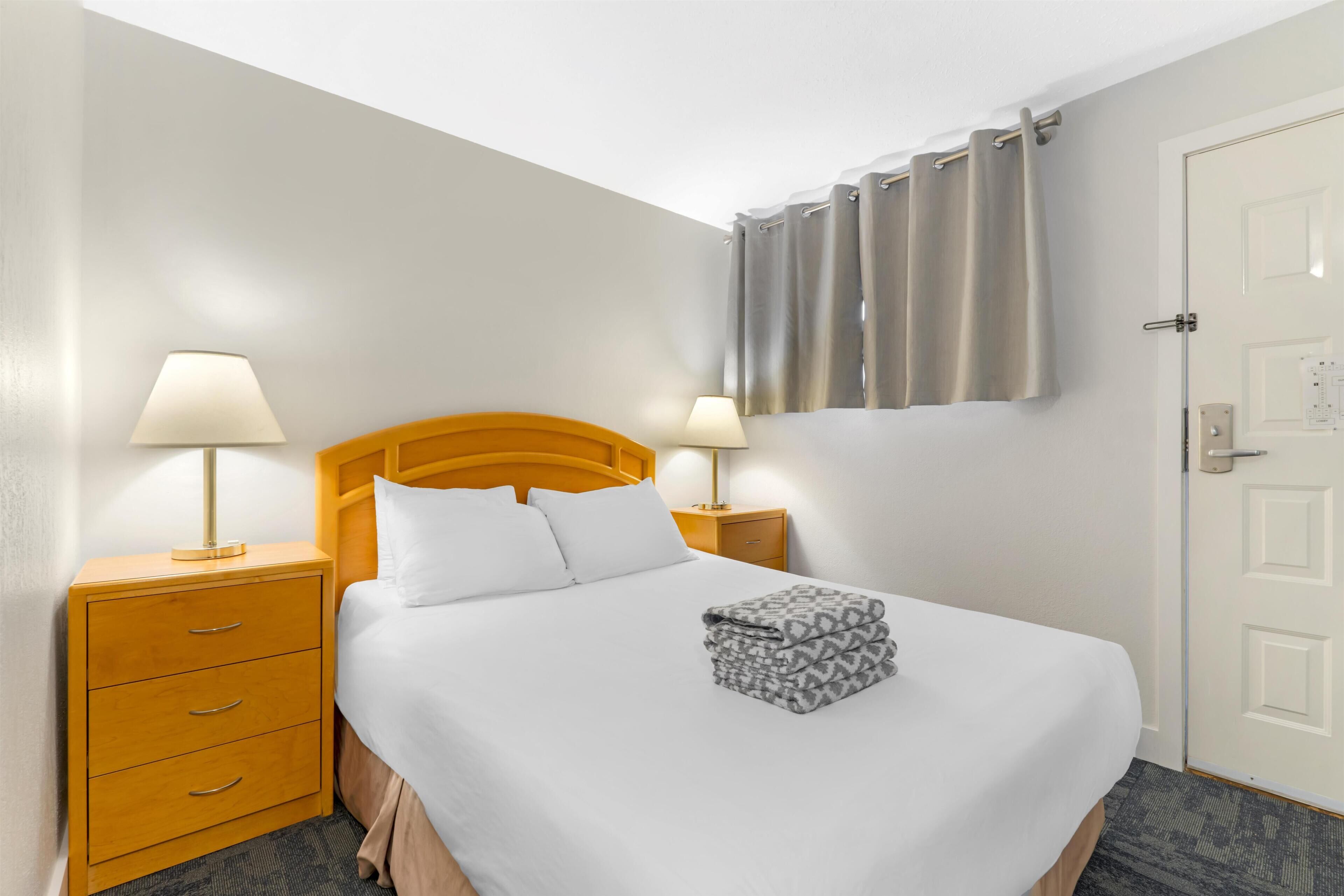 Una habitación de hotel con una cama, dos mesitas de noche con lámparas, un cabecero de madera, una puerta y una ventana con cortinas.