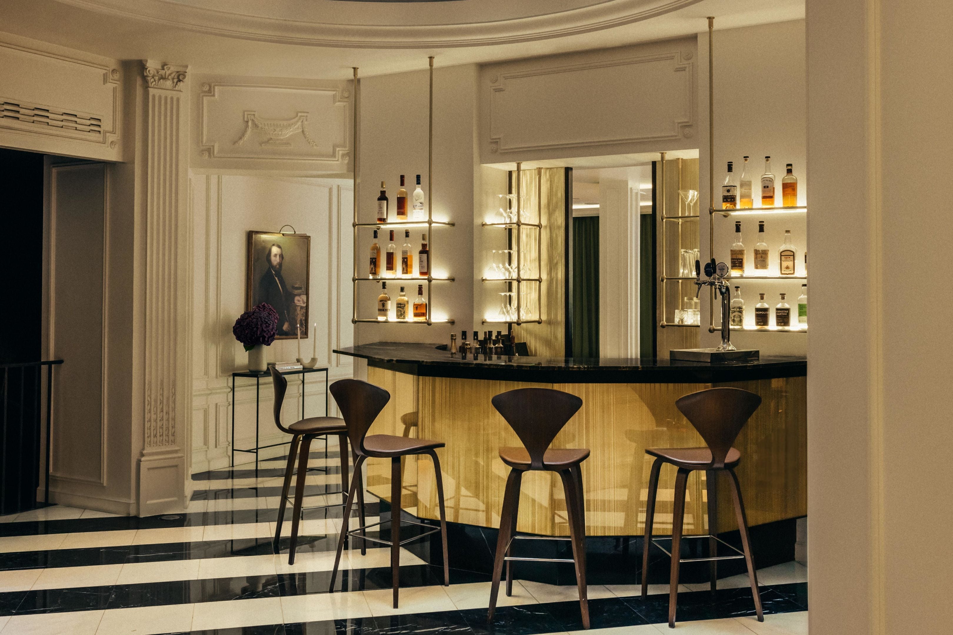 Uma área de bar com piso de mármore, balcão de madeira e três banquetas. Prateleiras com garrafas e copos atrás do balcão.
