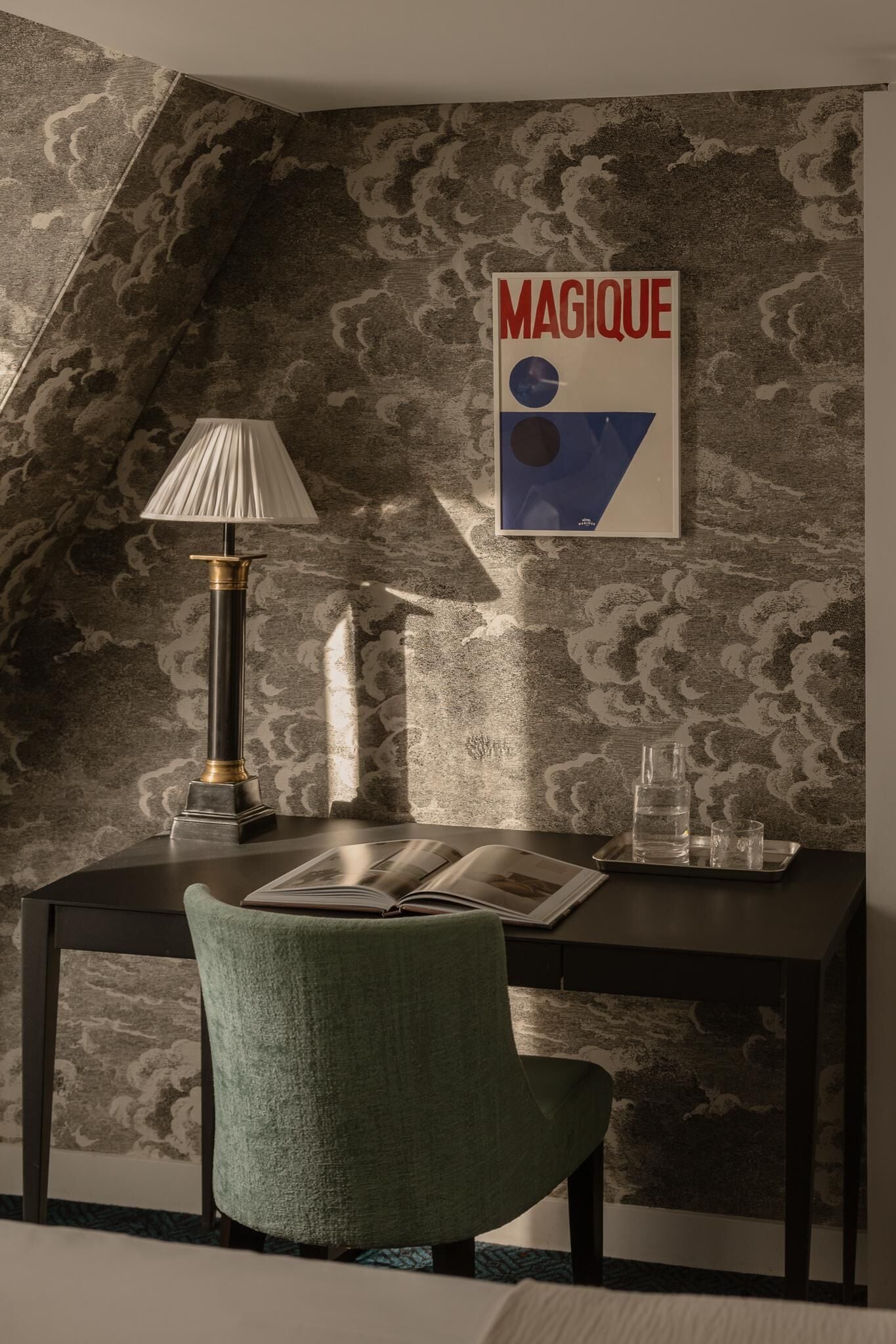 A corner room with a patterned wallpaper, a desk with a lamp, a chair, and a poster on the wall.