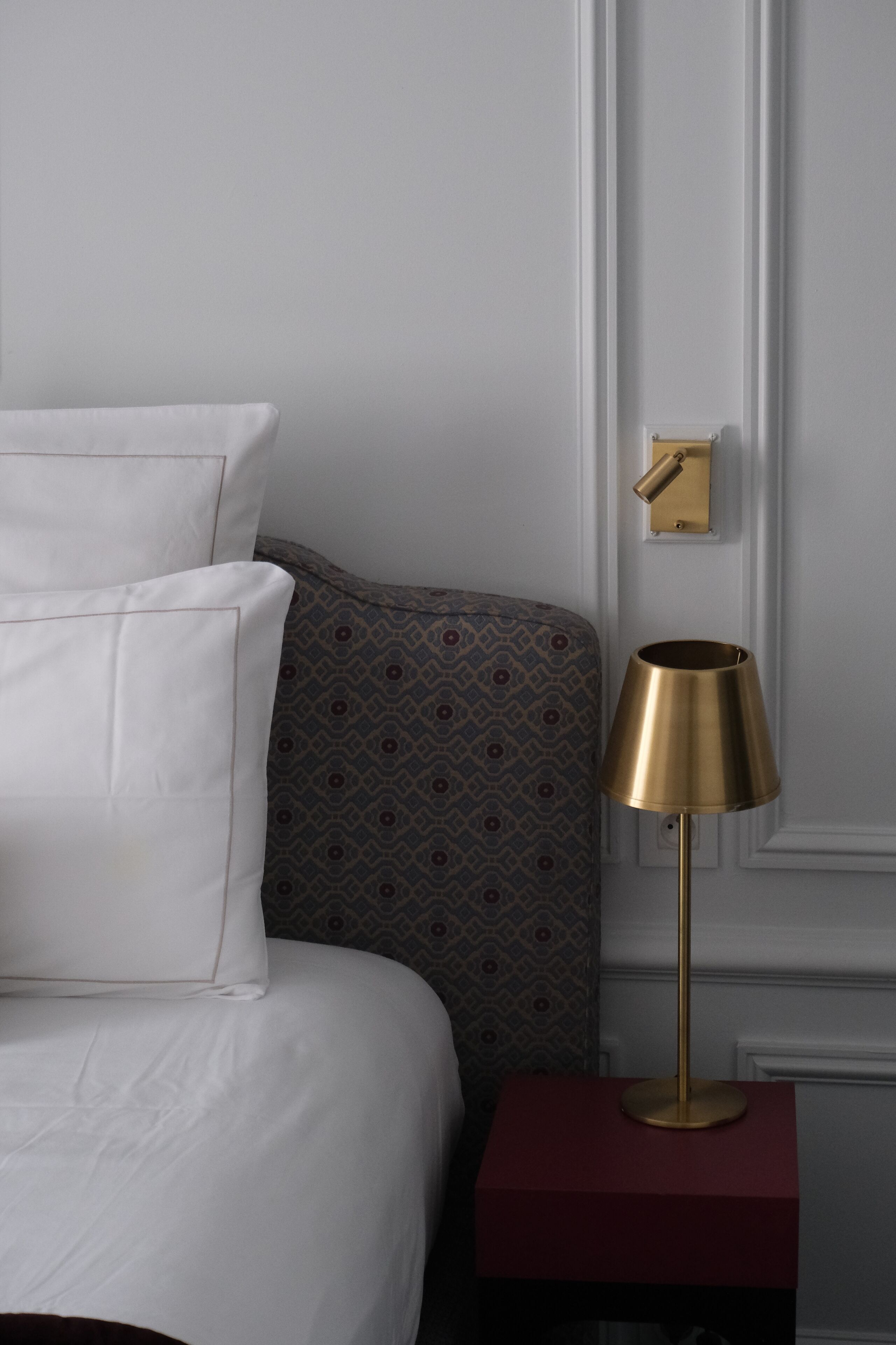 A neatly made bed with white linens and a patterned pillow, a gold lamp on a red nightstand, and a wall-mounted light switch.