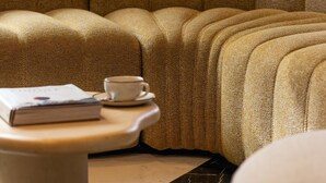 A corner sofa with a cushion, a small table with a cup and book, and a marble floor.