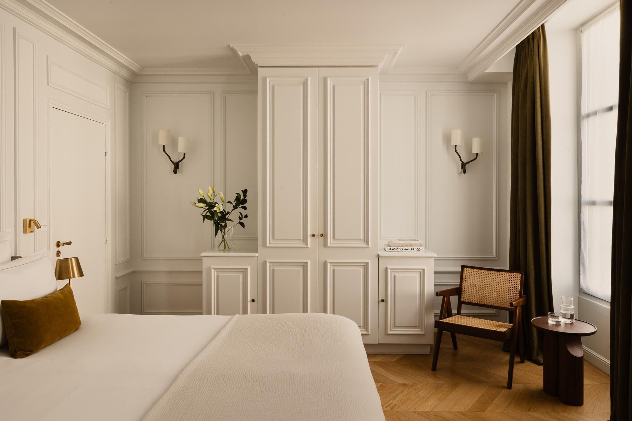 Une chambre d’hôtel avec un grand lit, une petite table, une chaise, un vase avec des fleurs et une armoire.