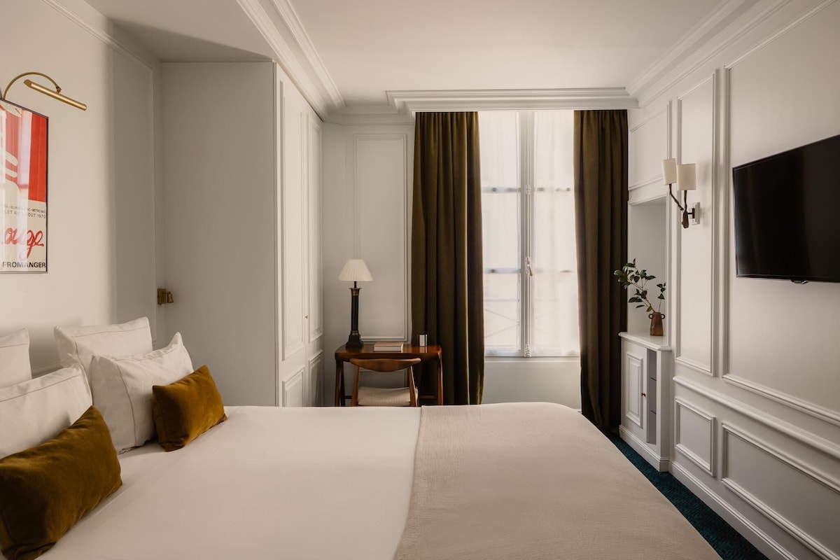 Une chambre d’hôtel avec un grand lit, un bureau avec une lampe, une télévision et une fenêtre avec des rideaux.