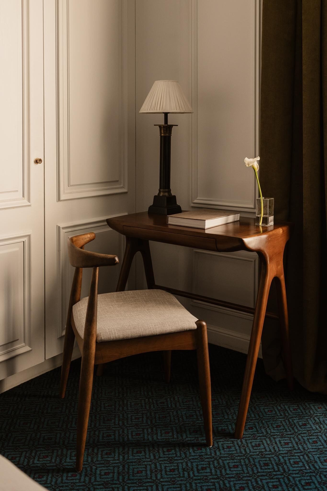 A room with a wooden desk, a chair, a lamp, a book, and a vase with a flower.