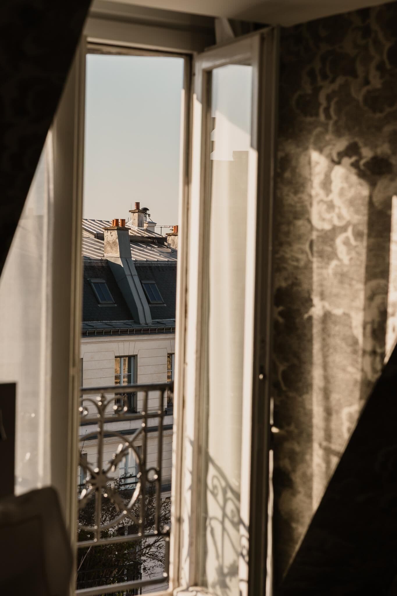 A view through a window with a metal railing, overlooking a building with a sloped roof and chimneys.