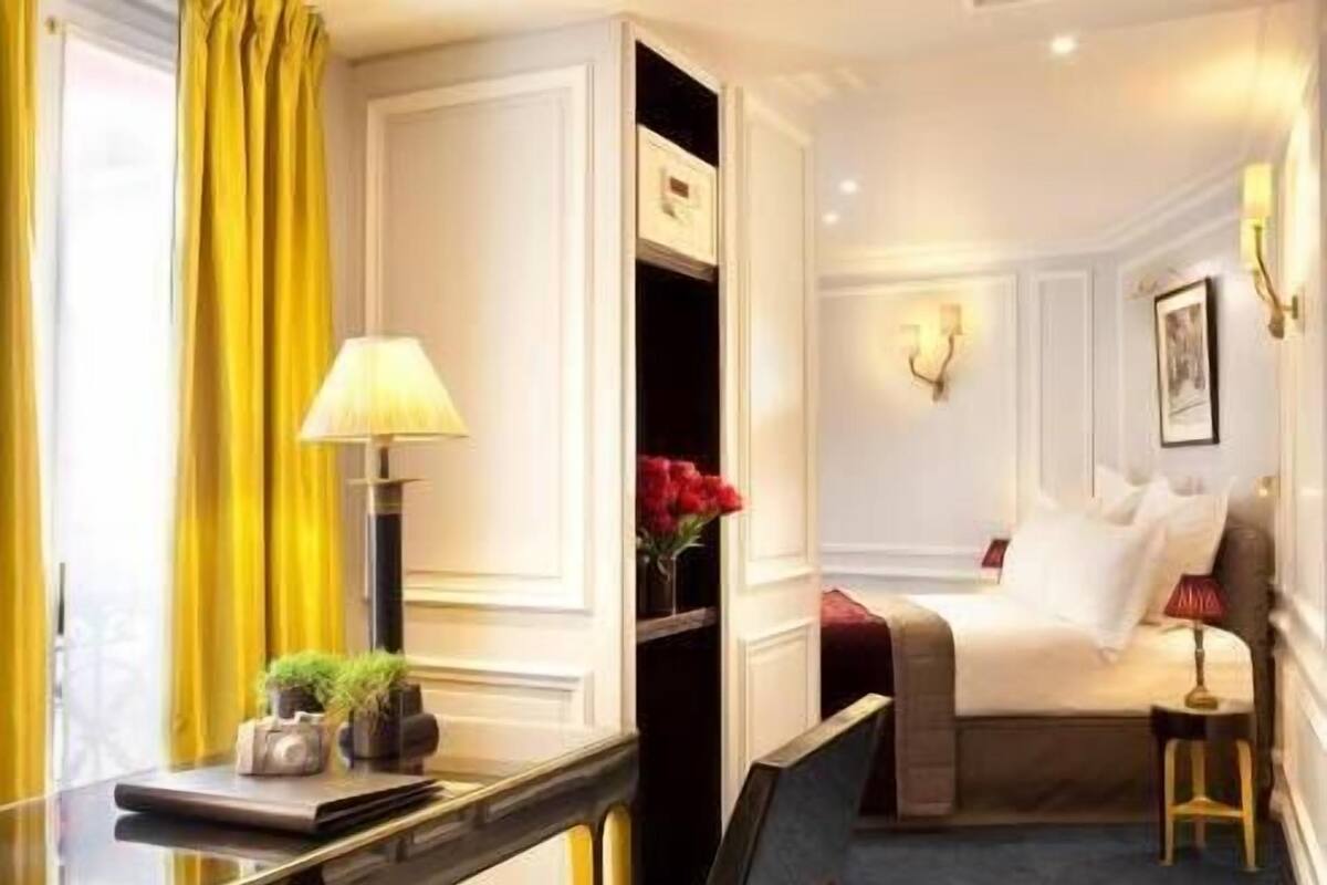 Une chambre d’hôtel avec un lit, un bureau doté d’une lampe. La chambre dispose d’une fenêtre avec des rideaux, d’une applique murale et d’une petite table sur laquelle est posé un vase de fleurs.