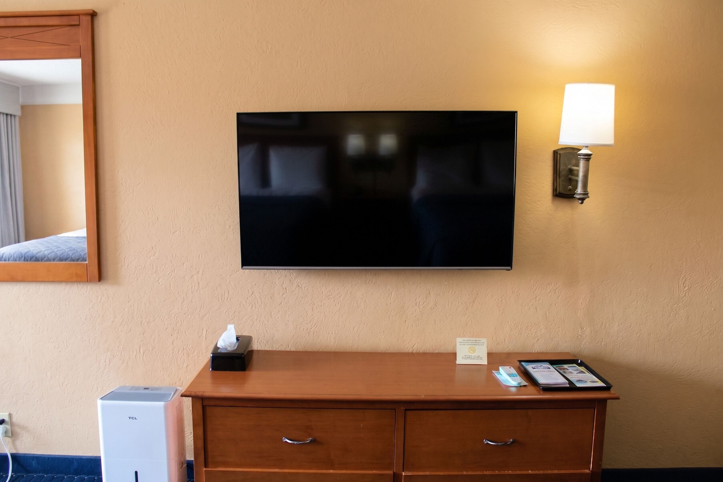 Ein Hotelzimmer mit Flachbildfernseher an der Wand, einer Holzkommode mit Schubladen, einer Papiertauschbox, einer Fernbedienung und einer Lampe.