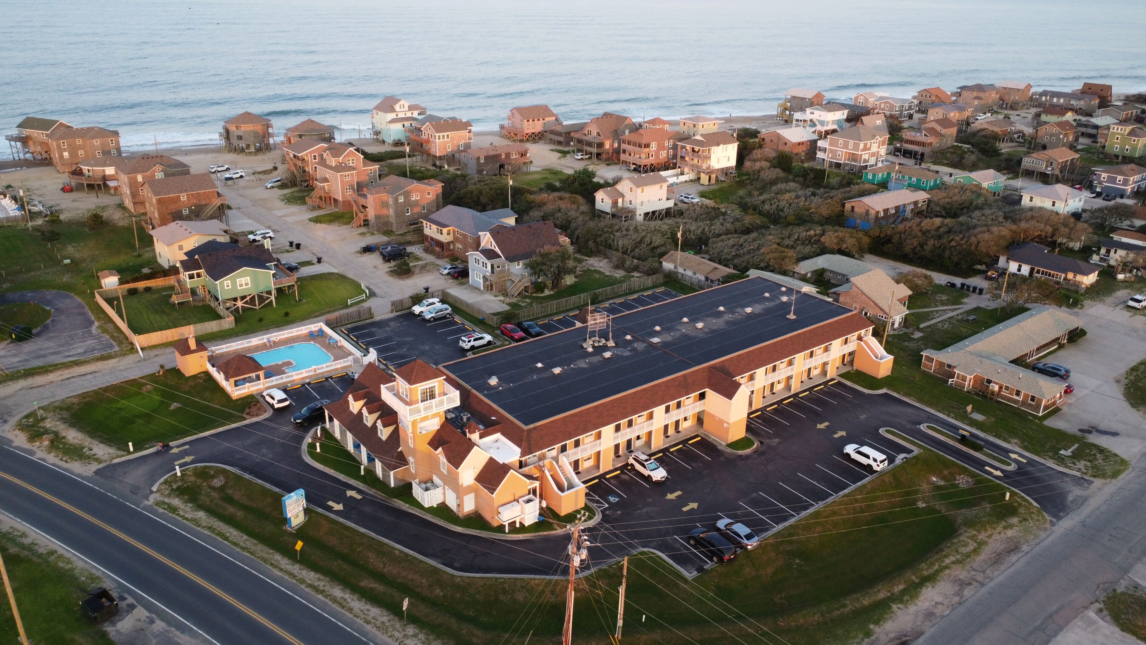 An aerial view of a coastal residential area with a large building featuring a parking lot and a swimming pool.