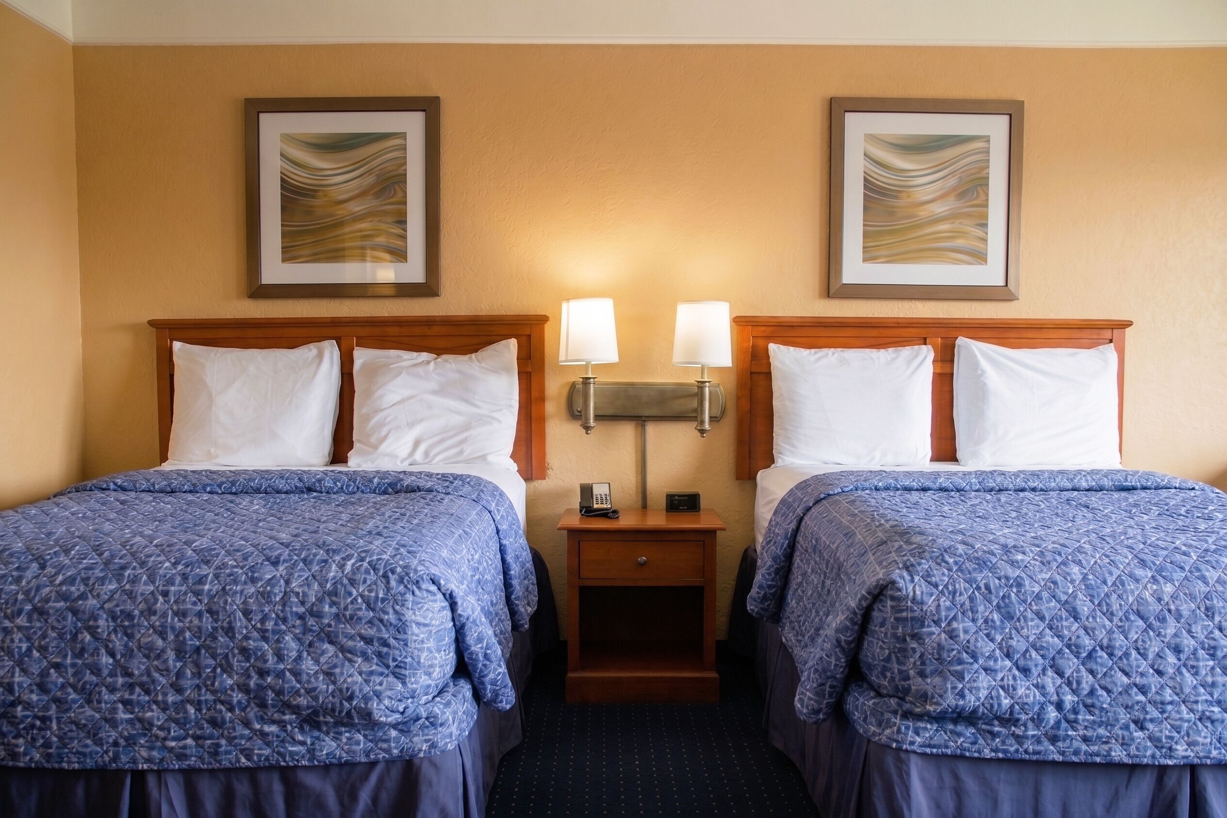 A hotel room with two beds, each with a blue quilted bedspread, a wooden headboard, and white pillows. There are two wall-mounted lamps on either side of a nightstand with a phone and a small table.