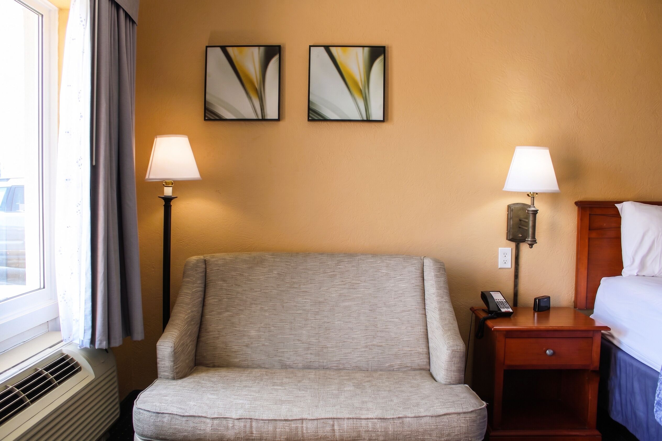 A hotel room with a single armchair, two bedside lamps, a bedside table with a phone and a small electronic device, and two abstract paintings on the wall.