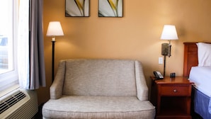 A hotel room with a single armchair, two bedside lamps, a bedside table with a phone and a small electronic device, and two abstract paintings on the wall.