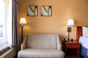 A hotel room with a single armchair, two bedside lamps, a bedside table with a phone and a small electronic device, and two abstract paintings on the wall.