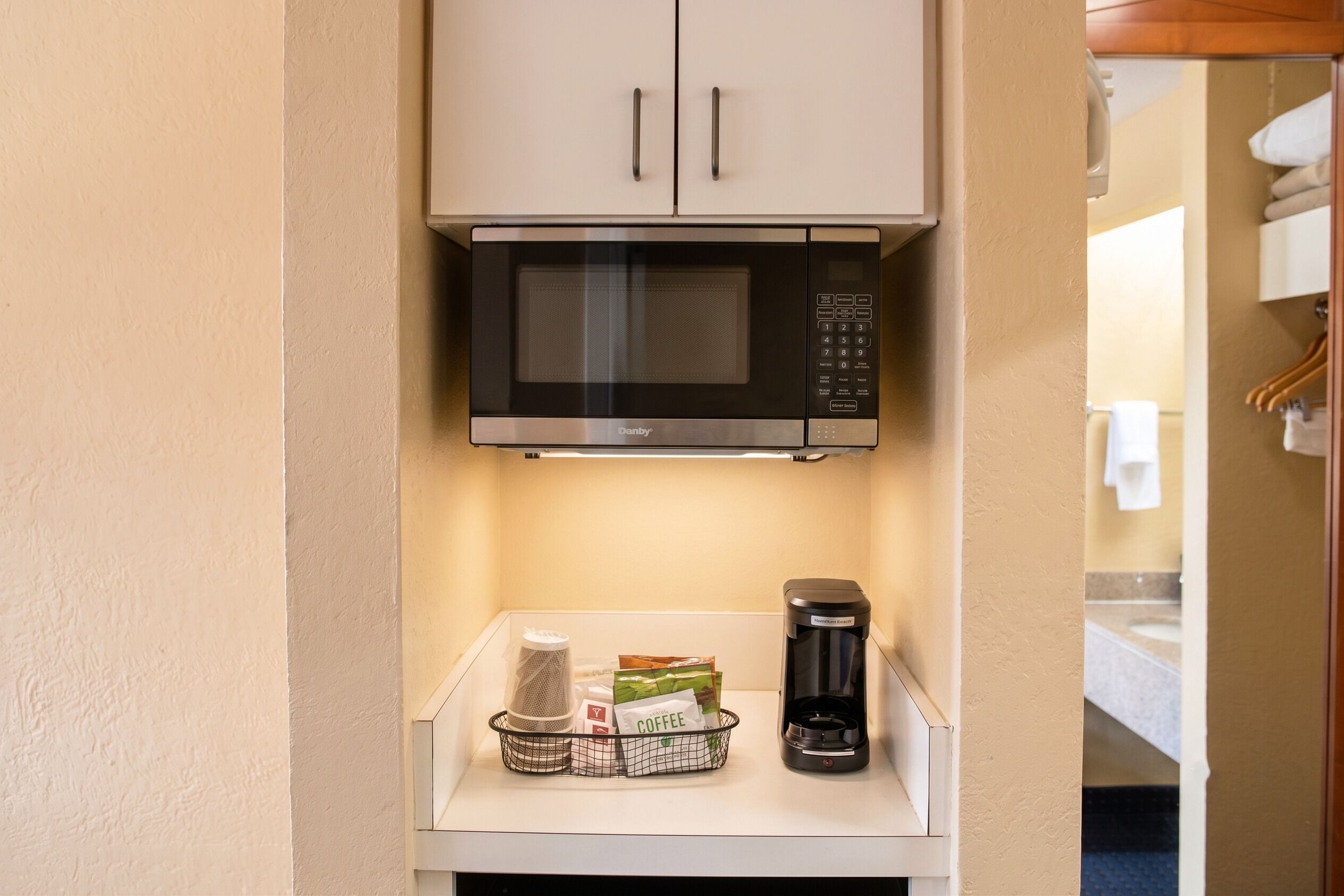 A microwave oven above a small table with a coffee maker and a basket containing a coffee bag and a mesh strainer.