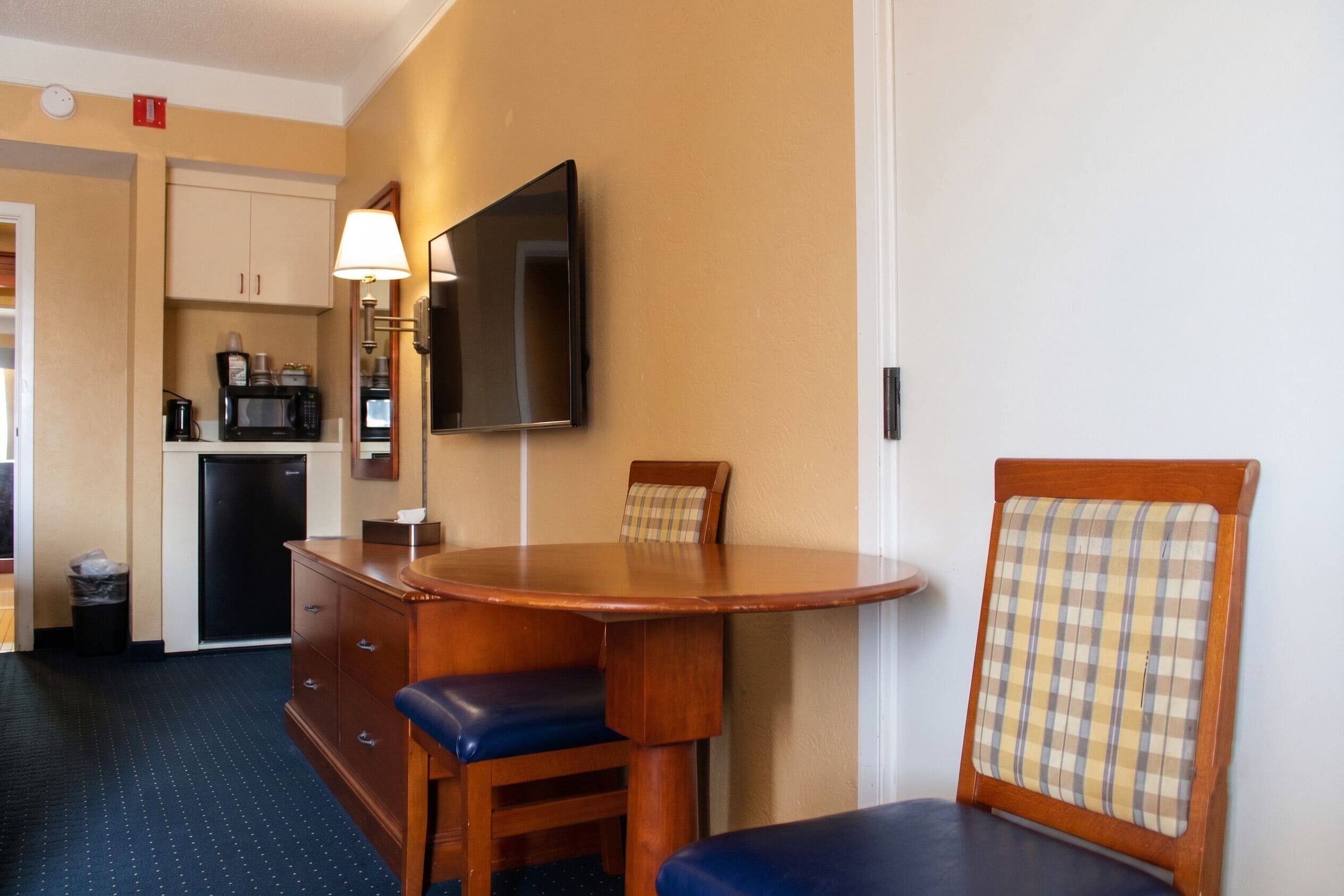 Habitación de hotel compacta con kitchenette, un televisor de pantalla plana montado en la pared, una mesa de madera con dos sillas y un pequeño cesto de basura.