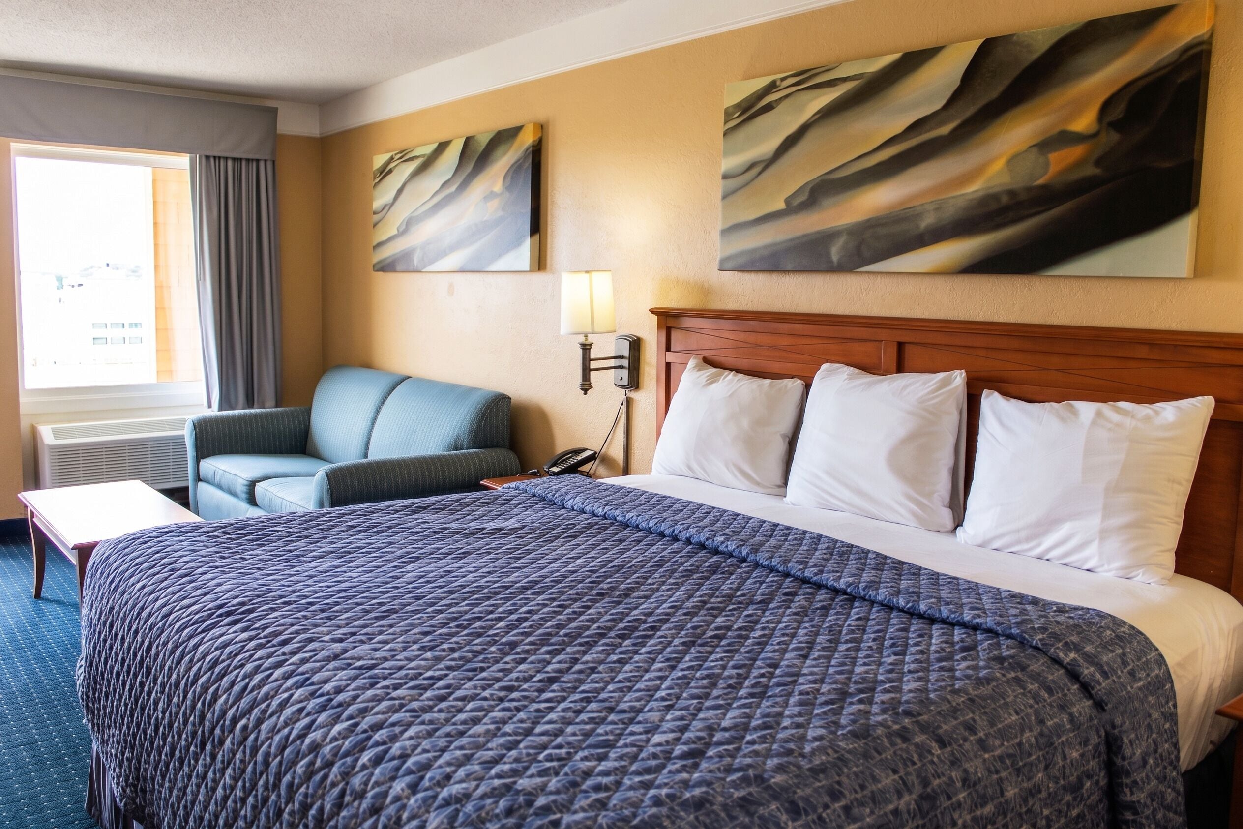 A hotel room with a large bed, a chair, a desk, and two paintings on the wall.