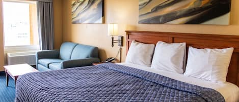 Quarto de hotel com uma cama grande, uma cadeira, uma escrivaninha e duas pinturas na parede.