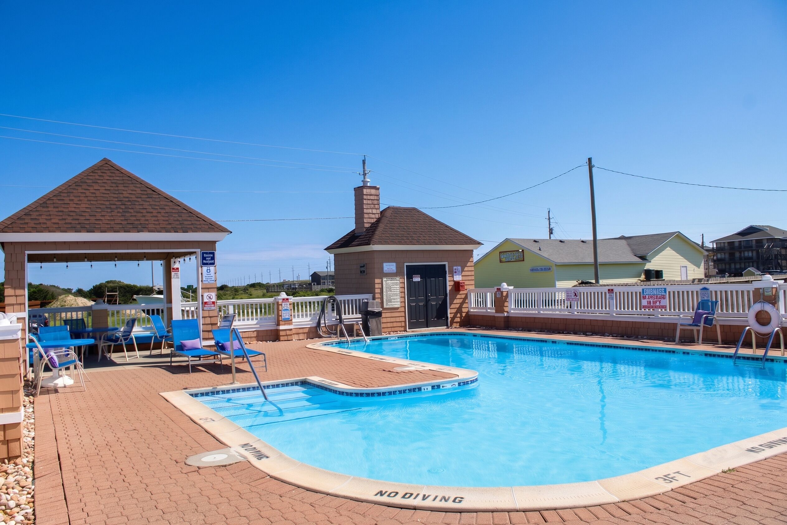 A swimming pool with lounge chairs and a covered area, surrounded by a fence and houses.