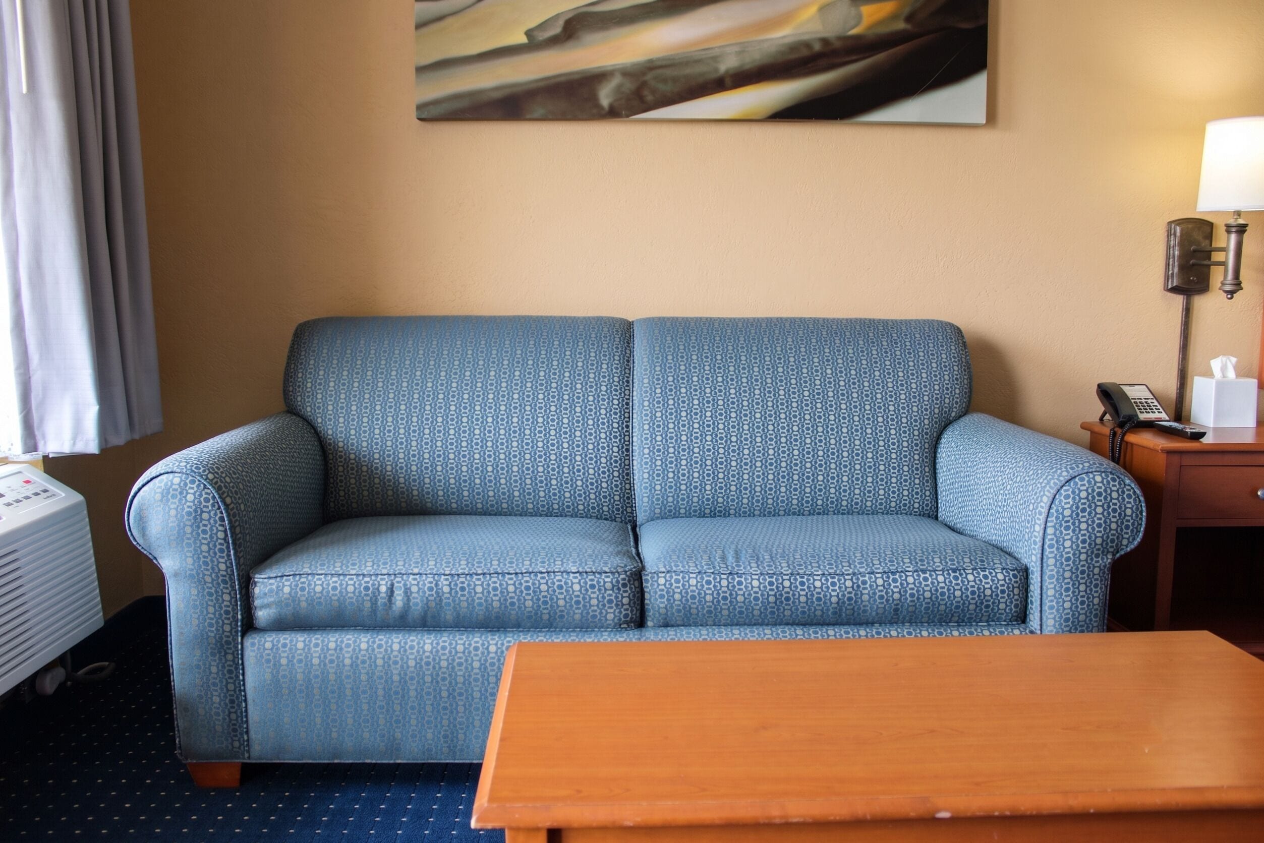 A hotel room with a blue patterned sofa, a wooden coffee table, a telephone, and a painting on the wall.
