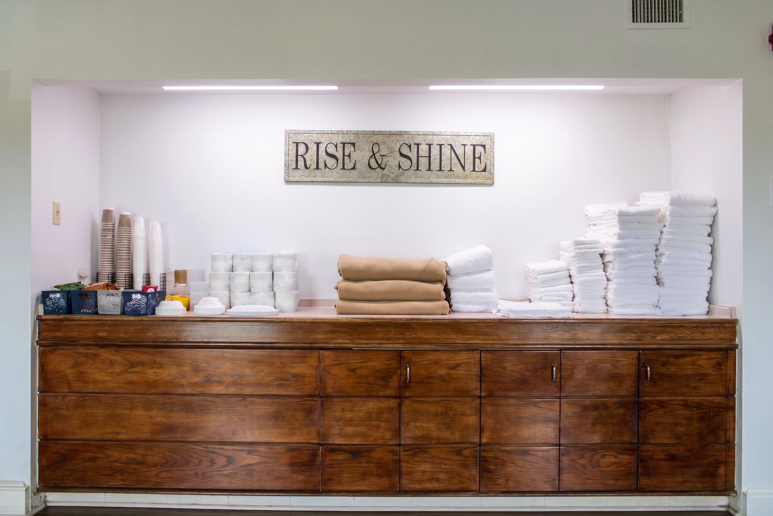 Uma área de recepção com um armário de madeira, uma placa com a inscrição 'RISE & SHINE' e pilhas de toalhas brancas.