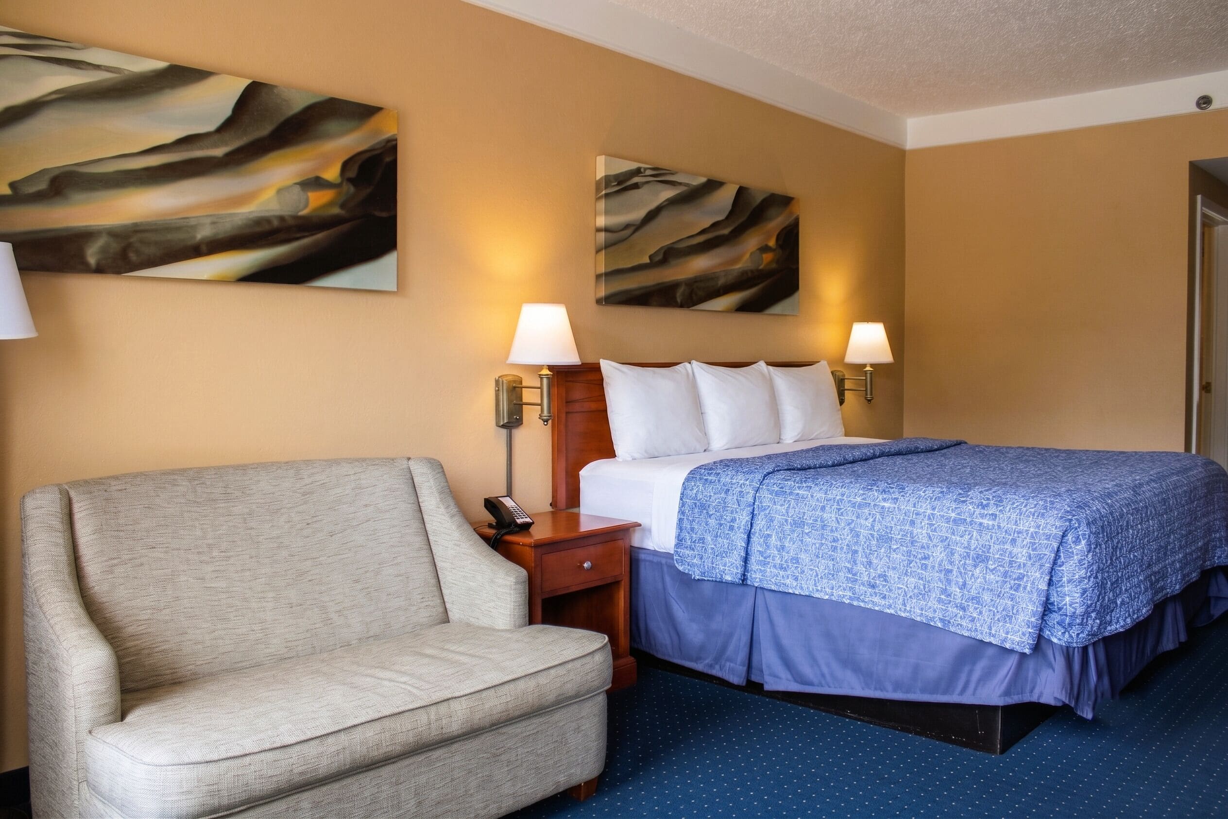 A hotel room with a bed, a sofa, two wall-mounted lamps, and two abstract paintings.