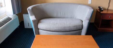 A room with a grey armchair, a wooden table, a white air conditioner, and a telephone on a wooden side table.