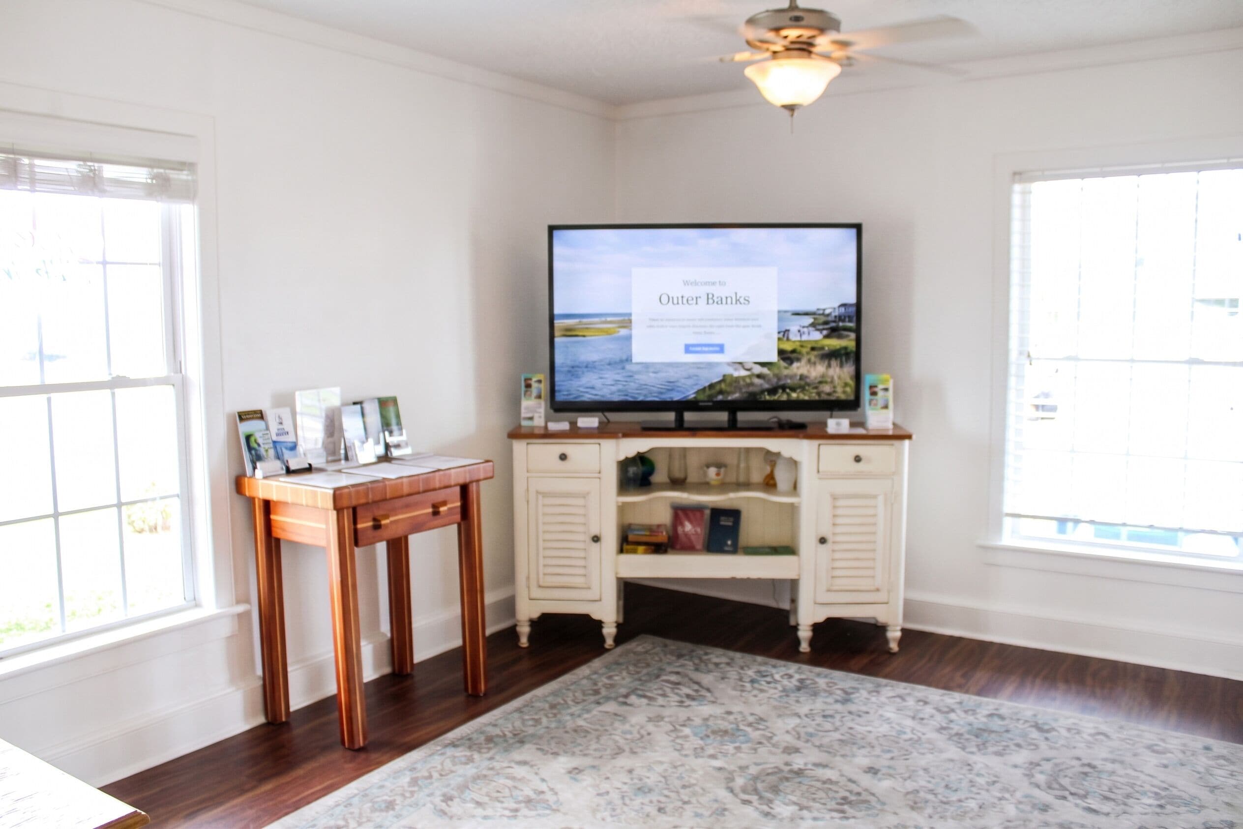 A room with a television displaying a welcome message, a wooden table with papers, a patterned rug, and a cabinet with items on top.