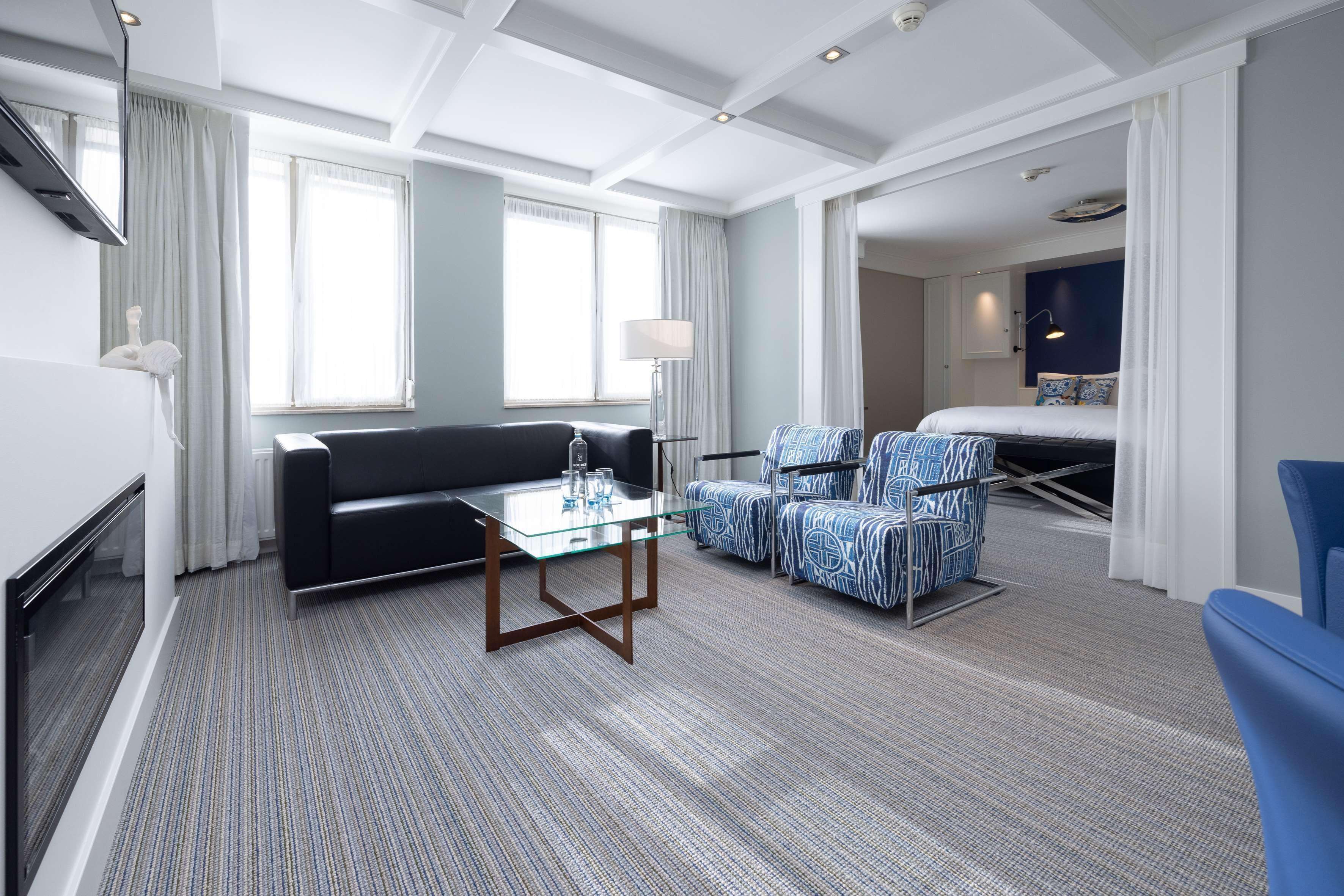 Habitación de hotel con sofá, dos sillones, una mesa de centro de cristal, televisor de pantalla plana, lámpara y cama con cabecero.