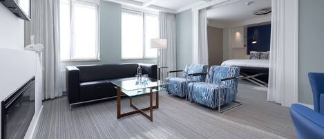 Habitación de hotel con sofá, dos sillones, una mesa de centro de cristal, televisor de pantalla plana, lámpara y cama con cabecero.