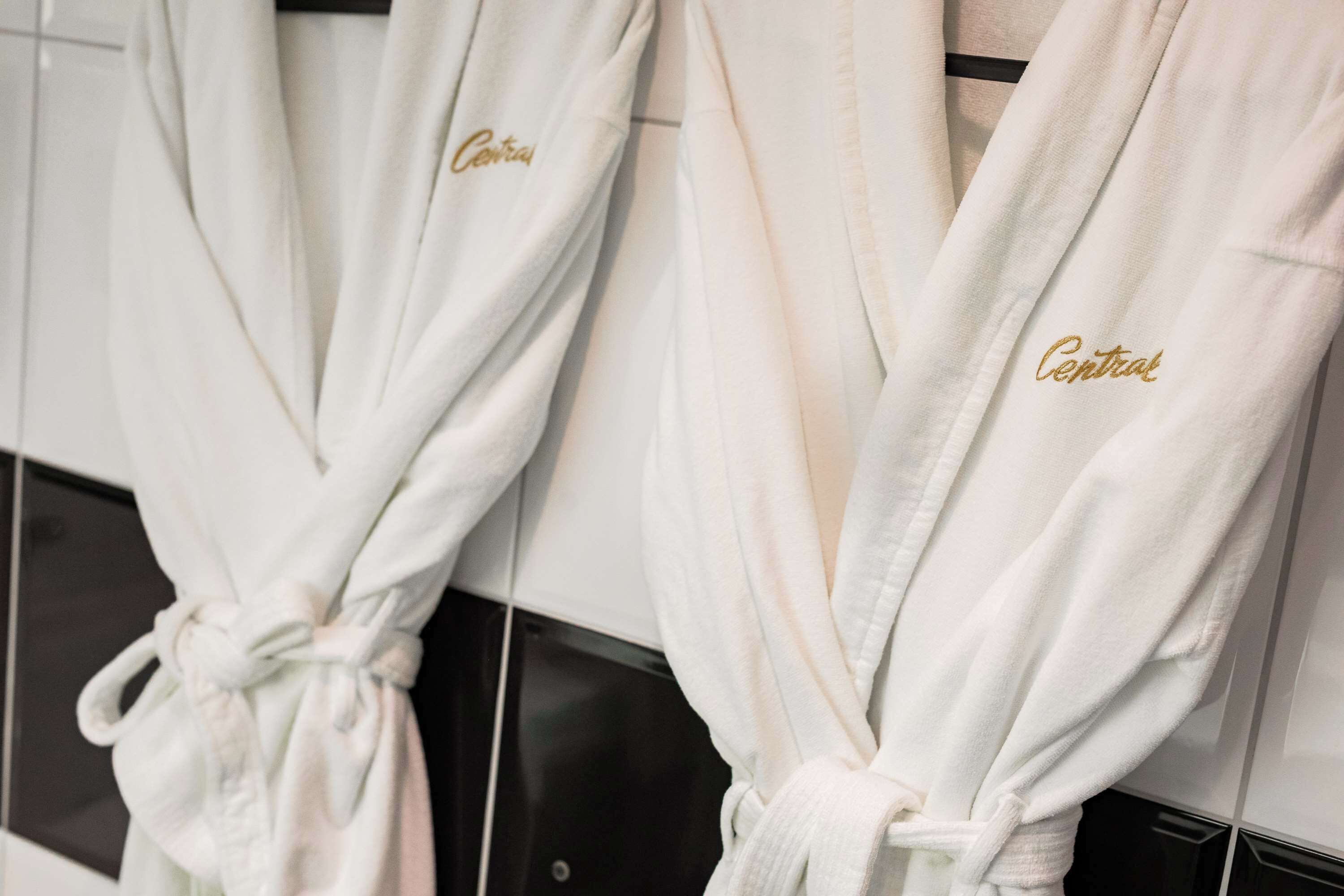 Dos batas blancas con el bordado 'Central' en dorado, colgadas en un perchero dentro de un baño.