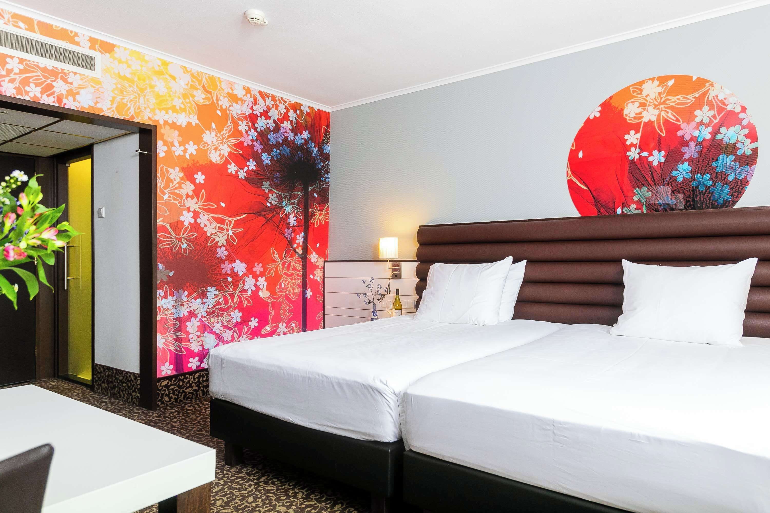 A hotel room with a double bed, a bedside table, a chair, a wall with floral wallpaper, and a door leading to another room.