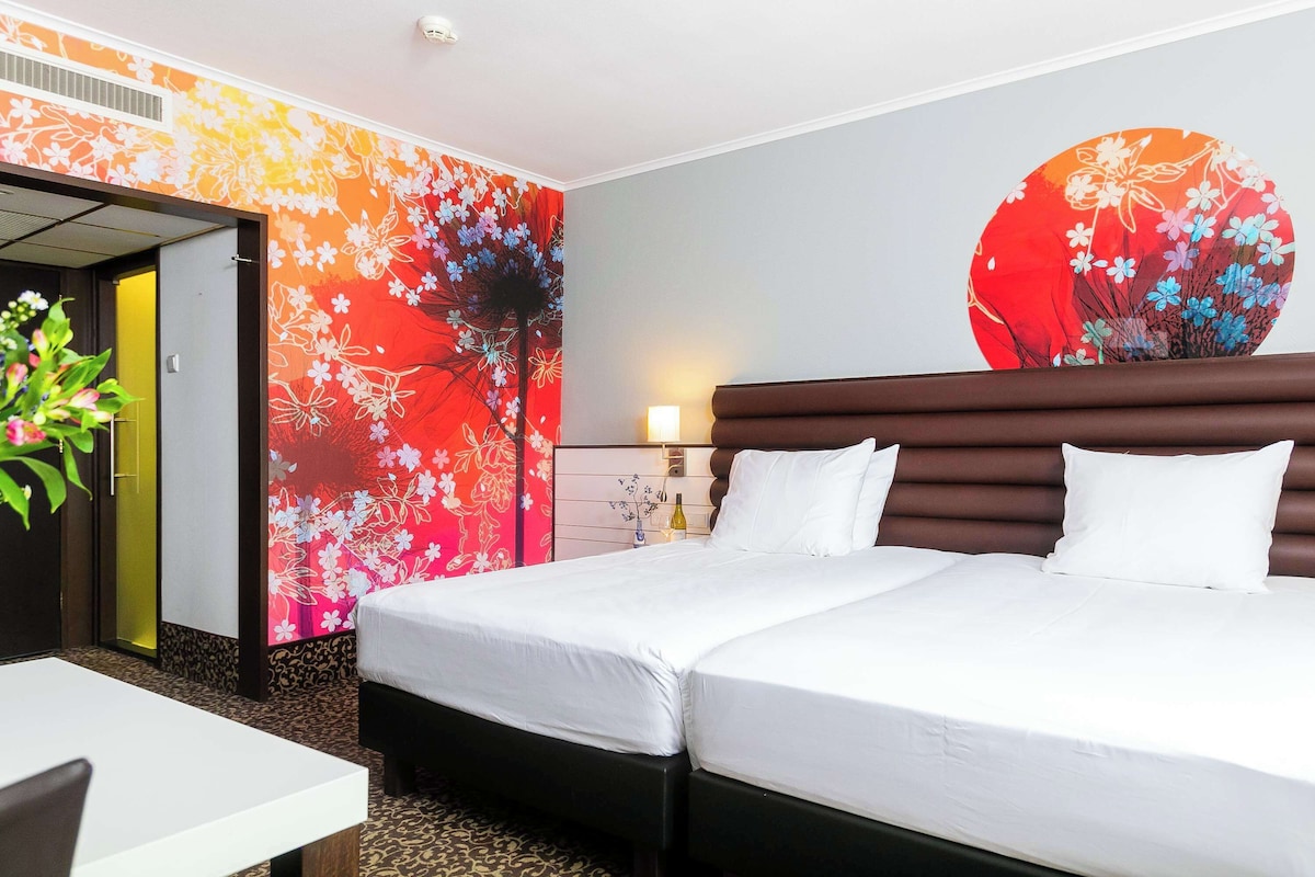 A hotel room with a double bed, a bedside table, a chair, a wall with floral wallpaper, and a door leading to another room.