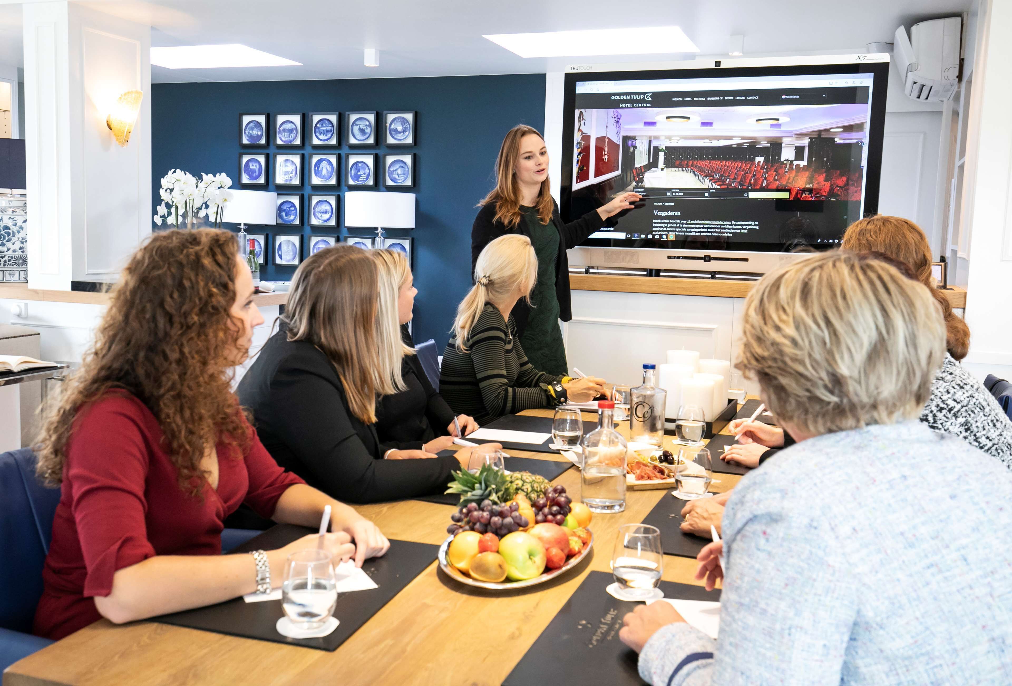 A meeting room with a presentation screen, a fruit platter, and attendees seated around a table.