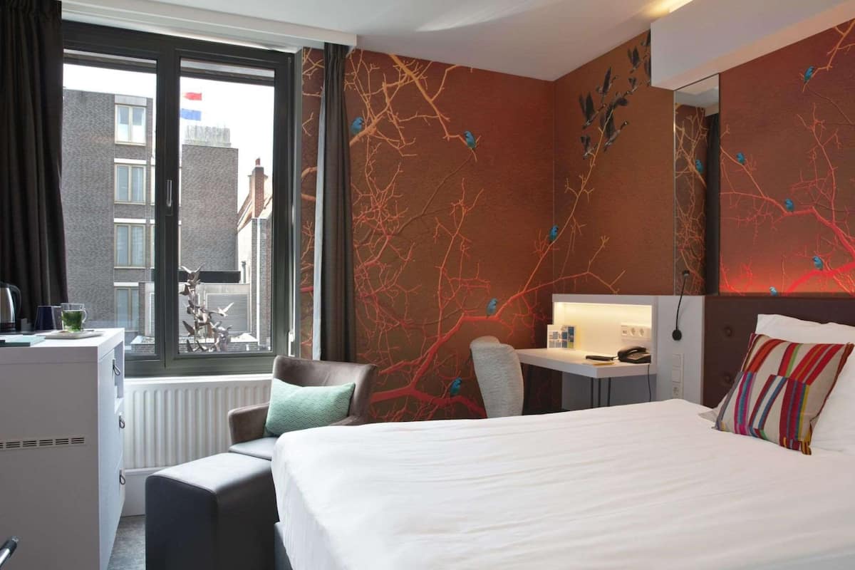 A hotel room with a bed, a desk, a chair, a radiator, and a window with a view of buildings.