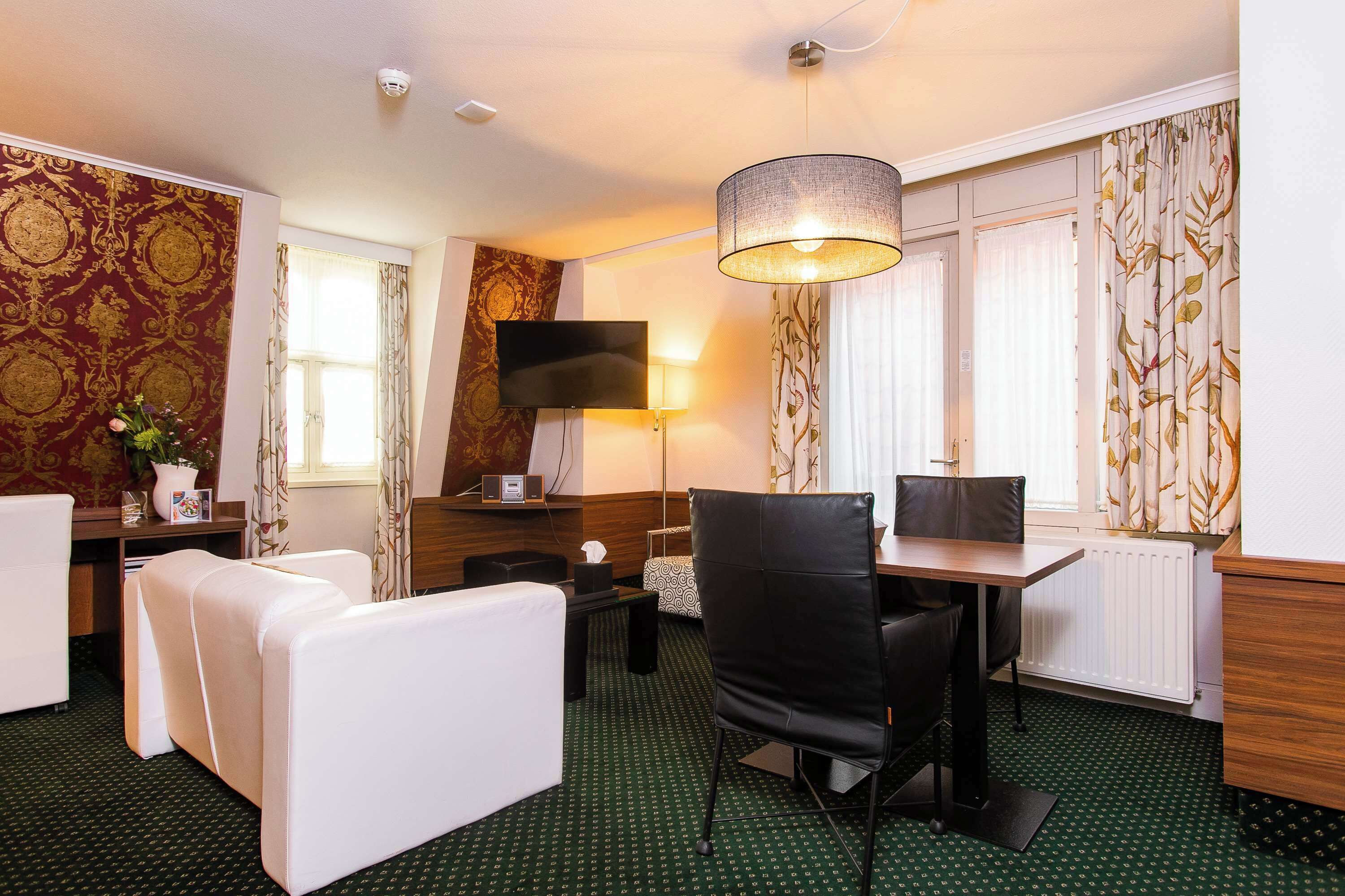 A hotel room with a green patterned carpet, a white sofa, a black chair, a wooden table, a TV, and a lamp.