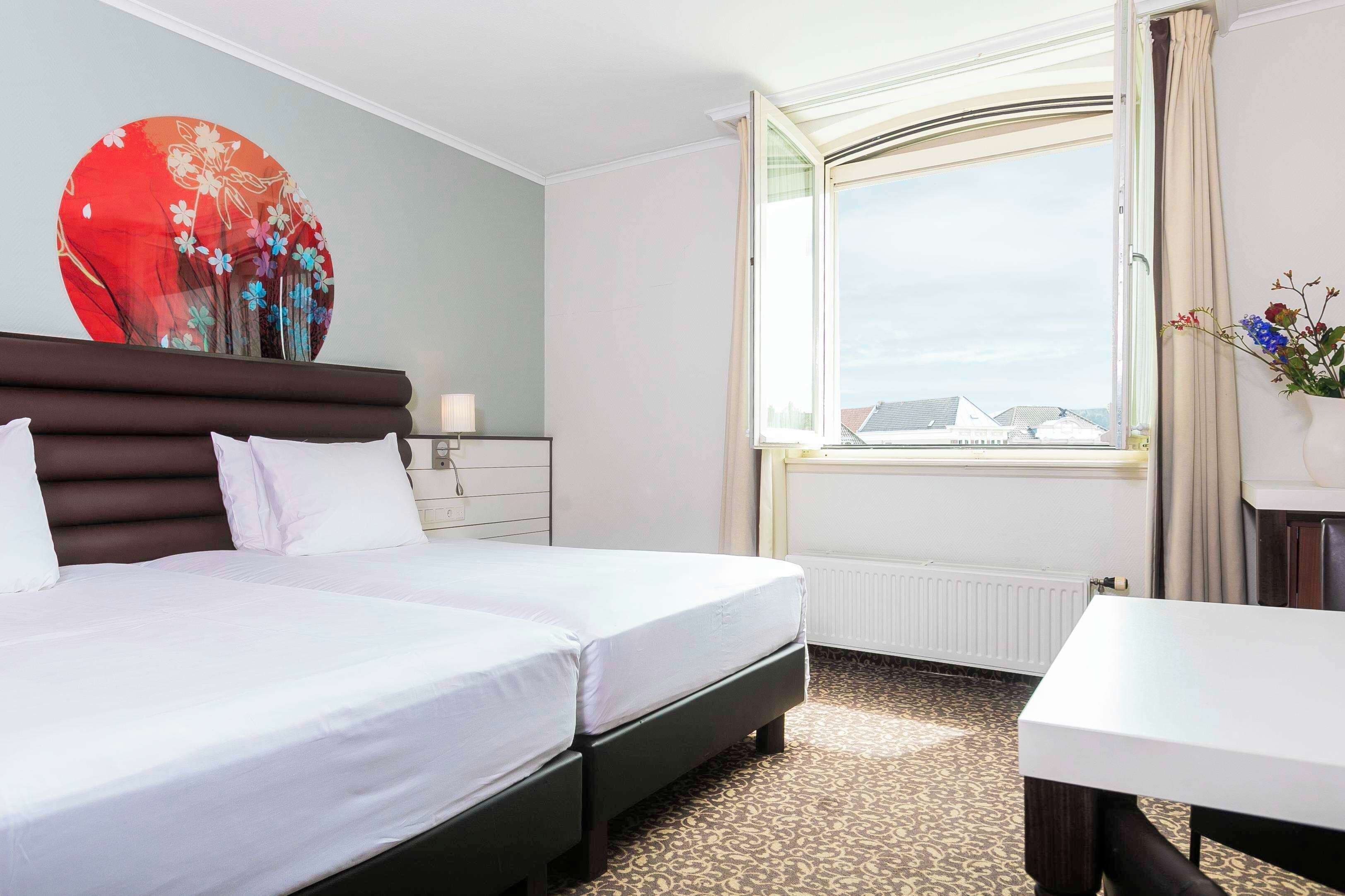 A hotel room with a bed, a bedside table, a window with curtains, and a wall decoration.