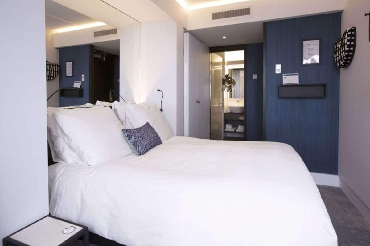 A modern hotel room with a large bed, a bedside table, a mirror, and a bathroom visible through an open door.