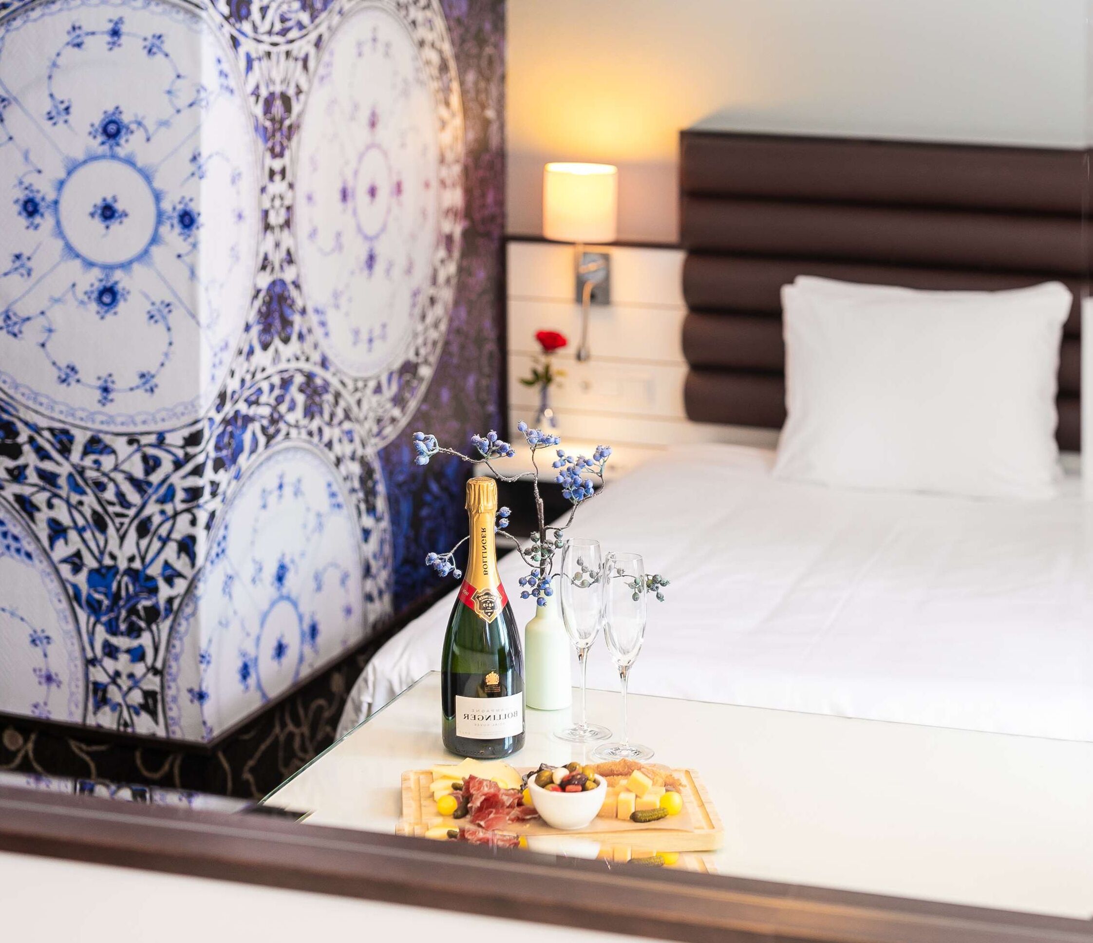 Habitación de hotel con una cama, una mesita de noche con una botella de champán, copas y una bandeja de frutas.