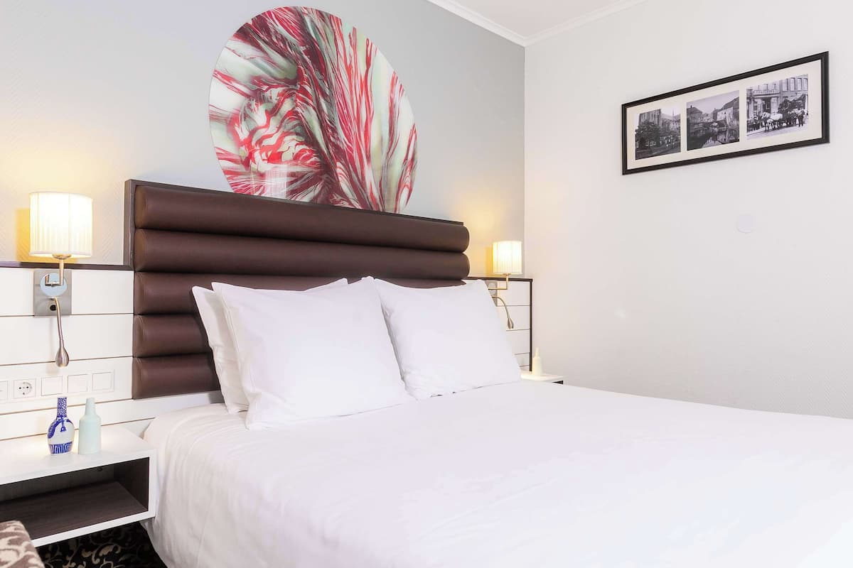 A hotel room with a large bed, a headboard, bedside lamps, a nightstand, and a wall with a large abstract painting and a framed photograph.