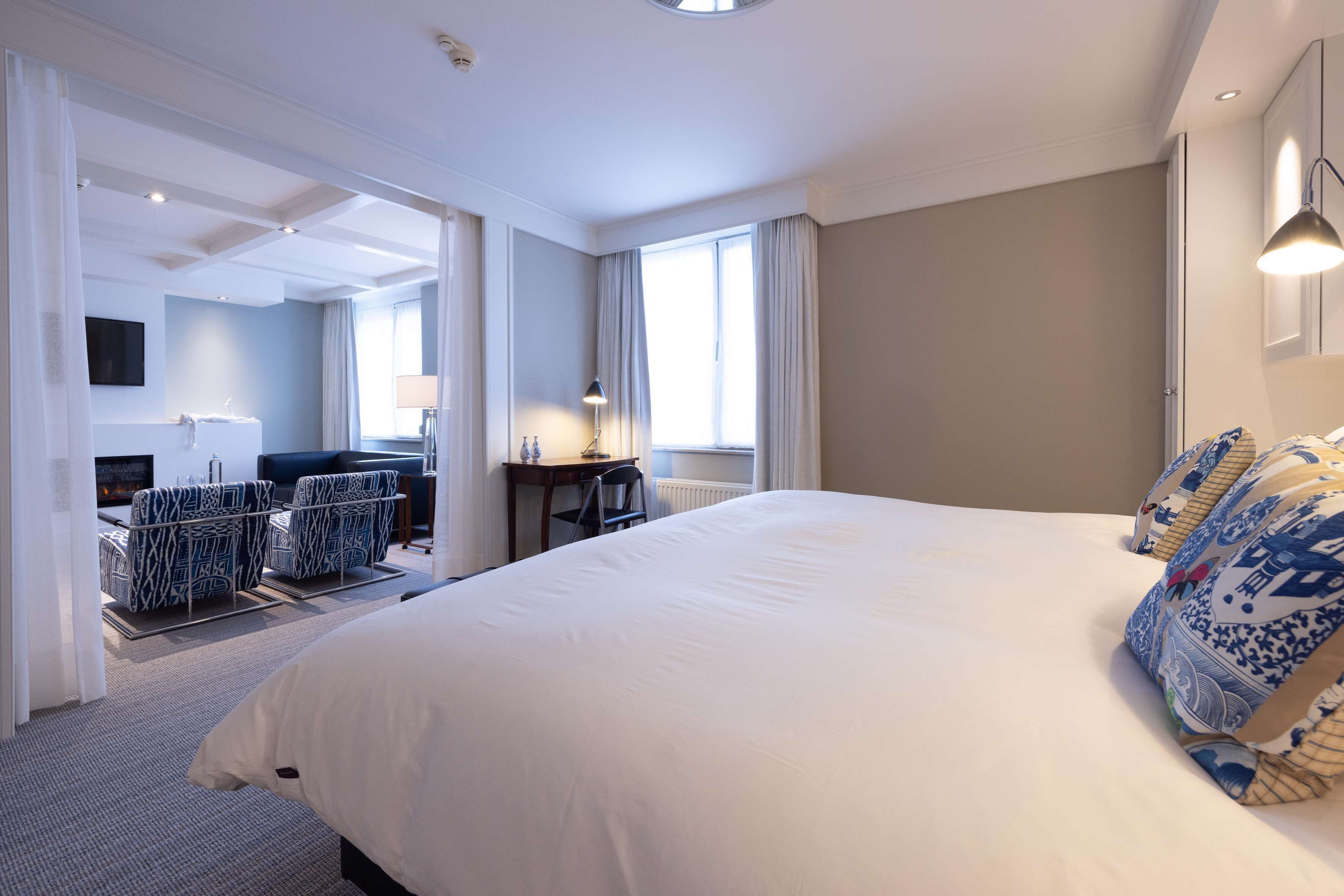 A hotel room with a large bed, a desk, two patterned chairs, a TV, and a window with curtains.