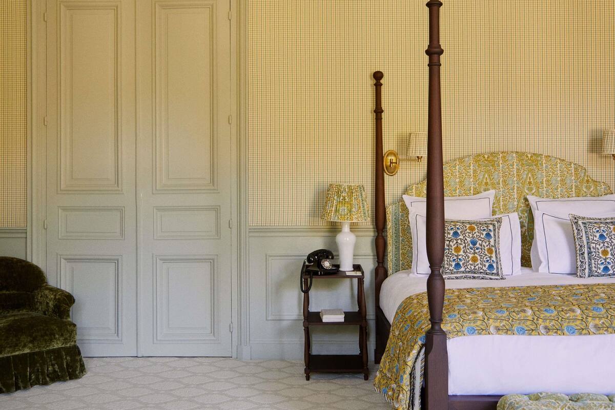 A four-poster bed with a floral bedspread, a bedside table with a lamp, and a dark armchair.