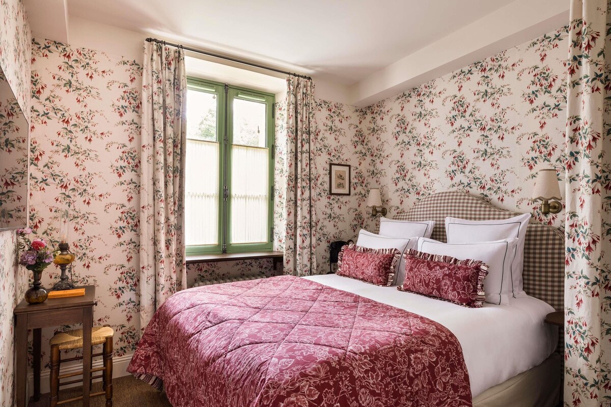 Une chambre à coucher avec un papier peint à motifs floraux, un lit avec une tête de lit à carreaux, une table de chevet et une fenêtre avec des rideaux.