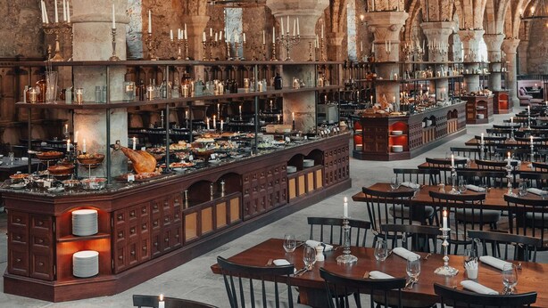 Uma sala de jantar ampla e bem iluminada, com tetos altos e arcos de pedra, com várias mesas e um bar central com diversos garrafas e copos.