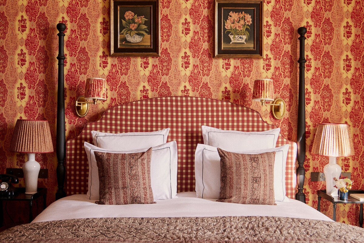 Een slaapkamer met een geruit hoofdeinde, een groot bed met veel kussens, een nachtkastje met een lamp, en een wandplank met een ingelijste foto.