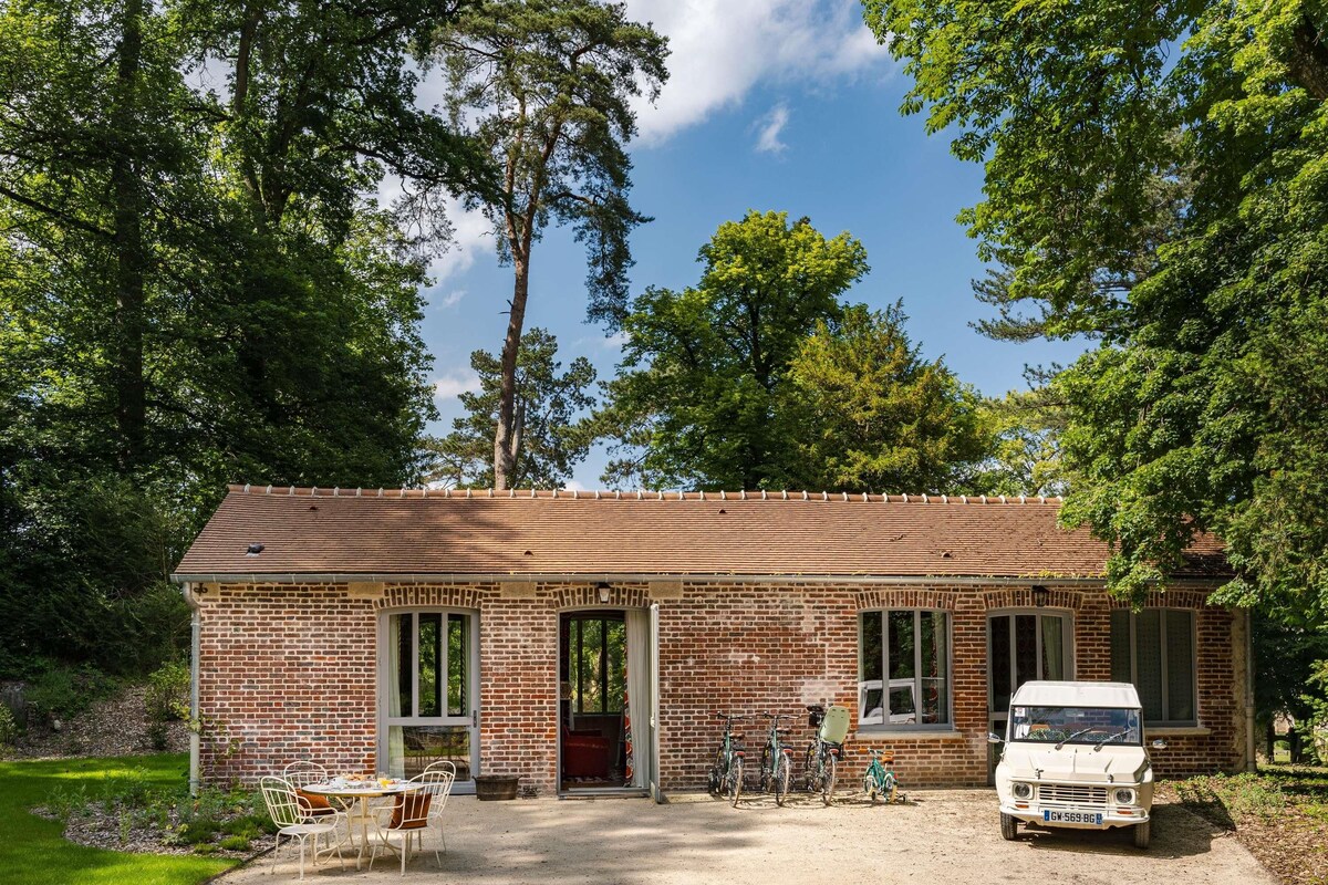 Un bâtiment en briques surmonté d’un toit en tuiles, avec une porte d’entrée et un petit porche doté de chaises. On peut voir des vélos garés à l’extérieur et un véhicule blanc devant.