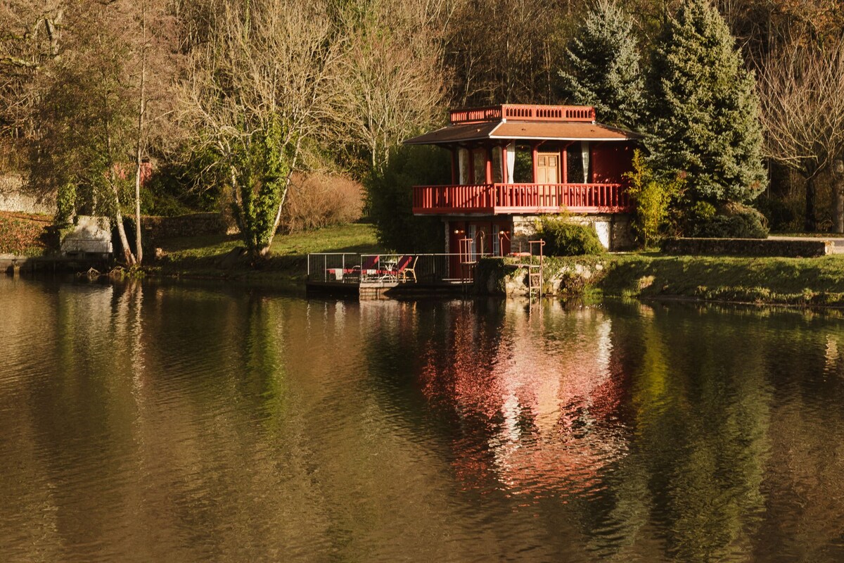 Een huis aan een meer met een rood balkon, omgeven door bomen en een heldere lucht.
