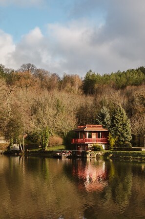 Une maison au bord d’un lac, dotée d’un balcon rouge, entourée d’arbres et sous un ciel dégagé.