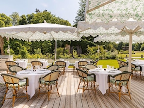An outdoor dining area with tables and chairs, surrounded by lush greenery and a stone wall.