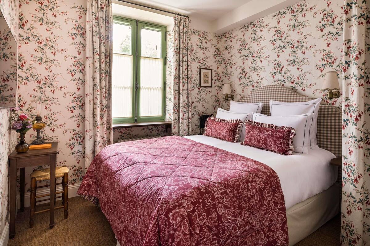 A bedroom with floral wallpaper, a bed with a checkered headboard, a nightstand, and a window with curtains.